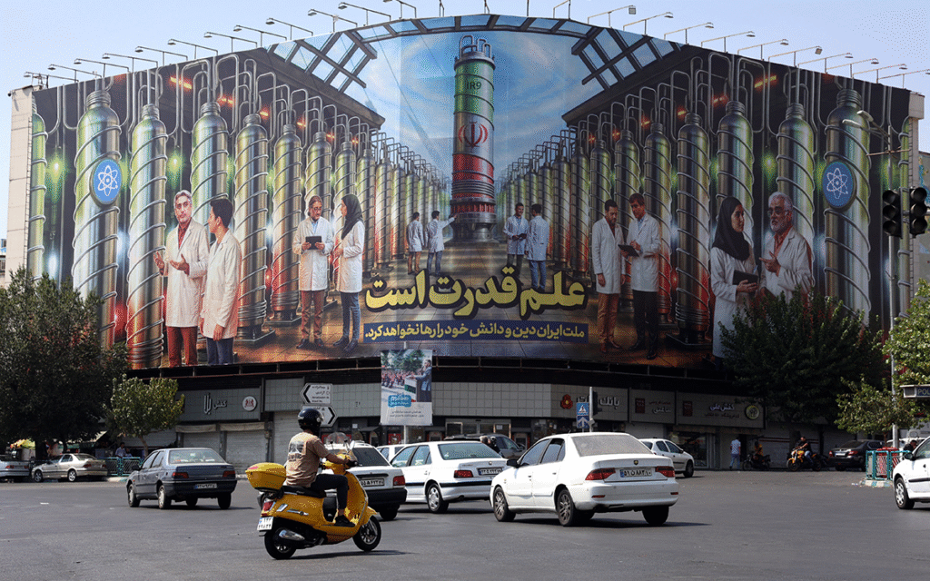 إيرانيون يقودون سياراتهم بجانب لوحة إعلانية تُظهر صورًا لعلماء نوويين وأجهزة الطرد المركزي (أ ف ب)