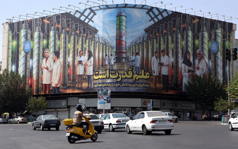 إيرانيون يقودون سياراتهم بجانب لوحة إعلانية تُظهر صورًا لعلماء نوويين وأجهزة الطرد المركزي (أ ف ب)