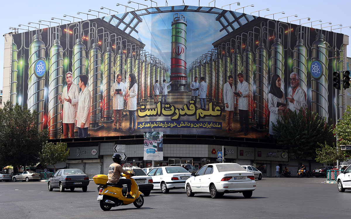 إيرانيون يقودون سياراتهم بجانب لوحة إعلانية تُظهر صورًا لعلماء نوويين وأجهزة الطرد المركزي (أ ف ب)